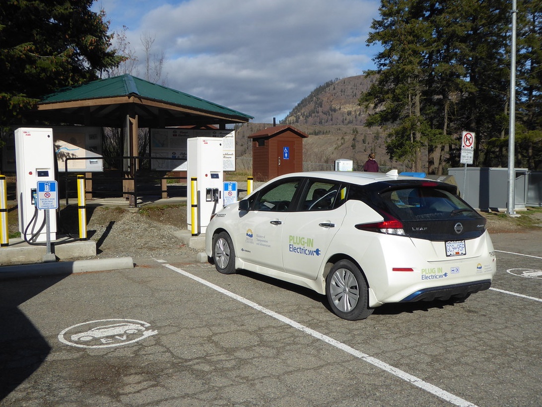 En vit elbil av modellen Nissan Leaf som laddas vid en offentlig snabbladdningsstation i ett bergslandskap.