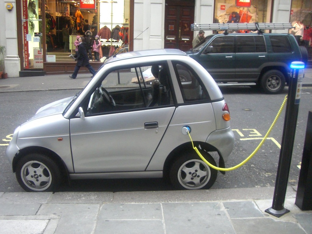 En kompakt vit elbil ansluten med en gul kabel till en blå laddbox på en stadsgata.